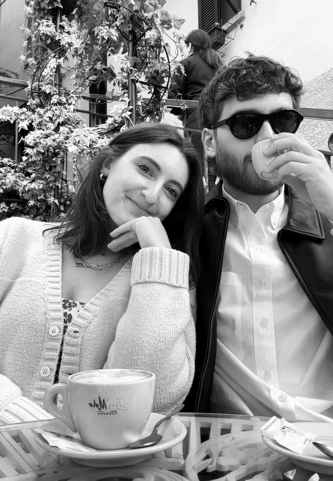 Sav and Drew enjoying coffee at an outdoor café in Italy. Sav has shoulder-length hair, a light sweater, and is smiling gently. Drew has curly hair, wears sunglasses, a white shirt, and a leather jacket, and is drinking from a cup.