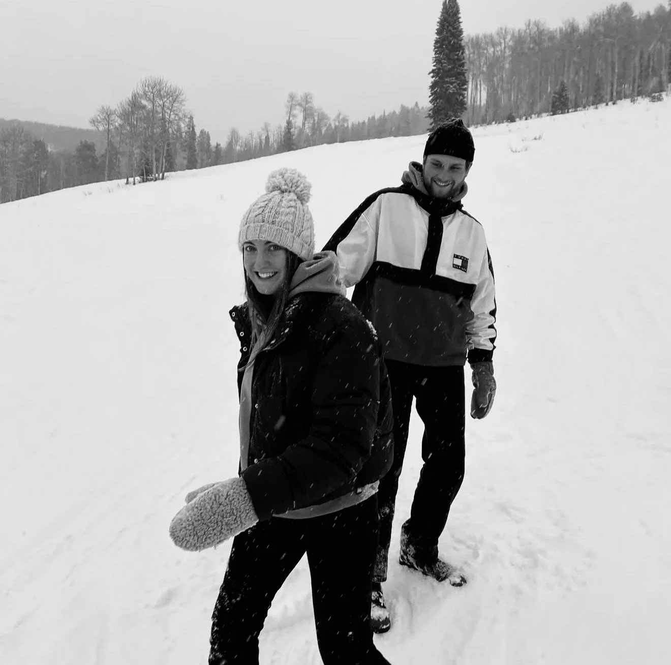 Drew and Sav smiling and standing in the snow on a winter day, with trees and mountains in the background.
