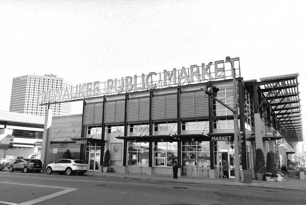 Black and white photo of a building with a sign reading Milwaukee Public Market, cars parked in front, and a traffic light in the foreground.