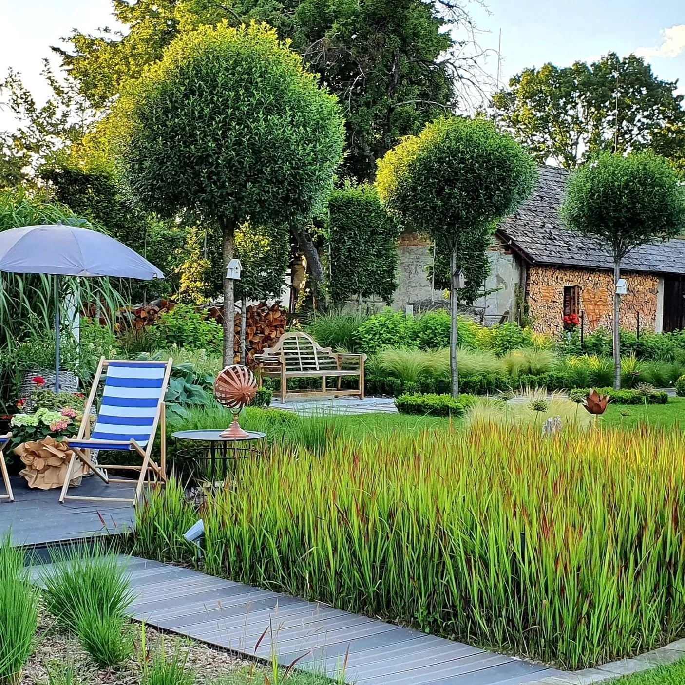 A lush garden with trimmed trees, flower beds, a lounging chair with an umbrella, and a wooden bench. There is a rustic stone house in the background and decorative garden ornaments, with a wooden pathway winding through the greenery.