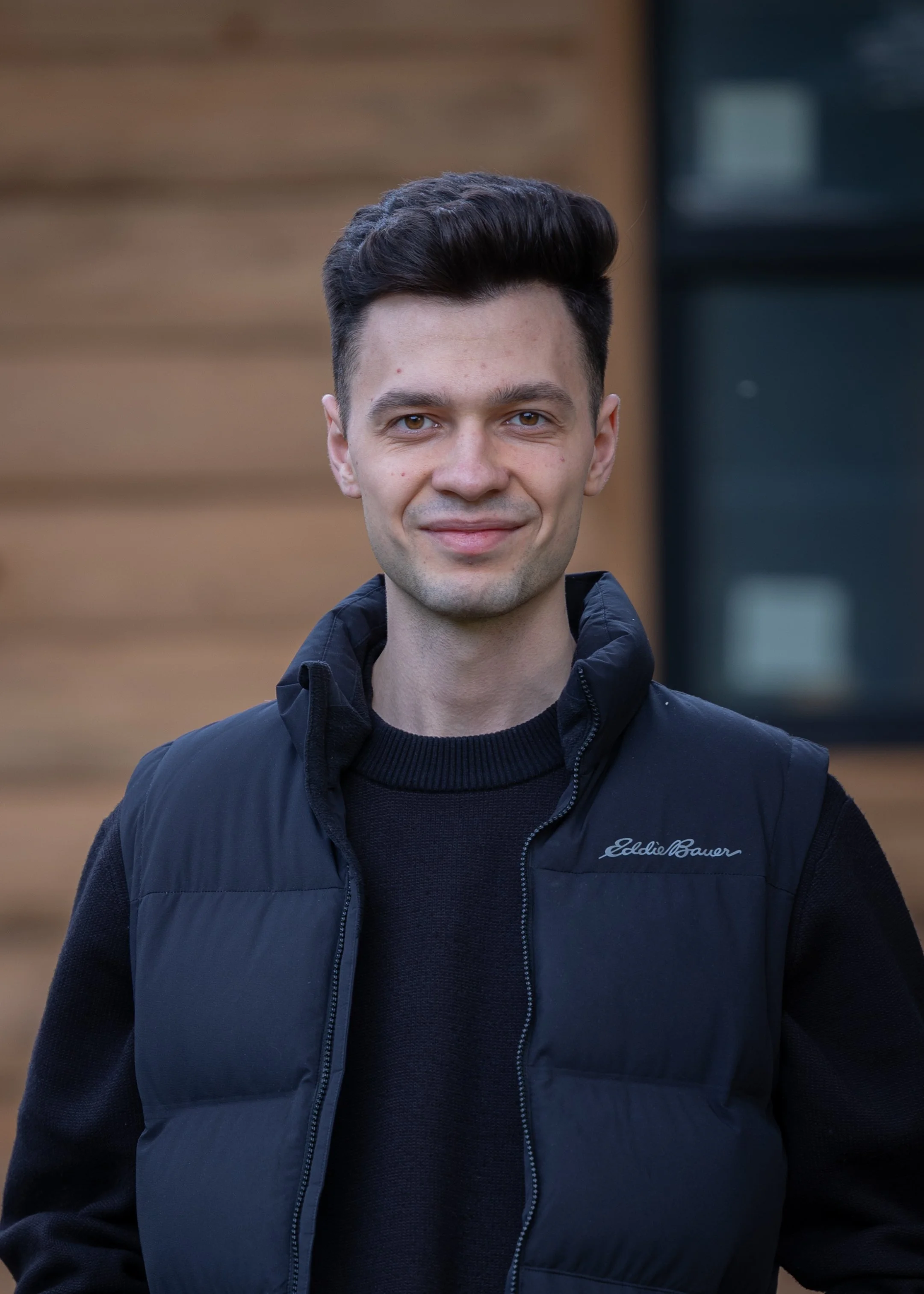 A young man with dark hair smiling outdoors, wearing a black sleeveless jacket over a dark sweater, with a blurred wooden building background.