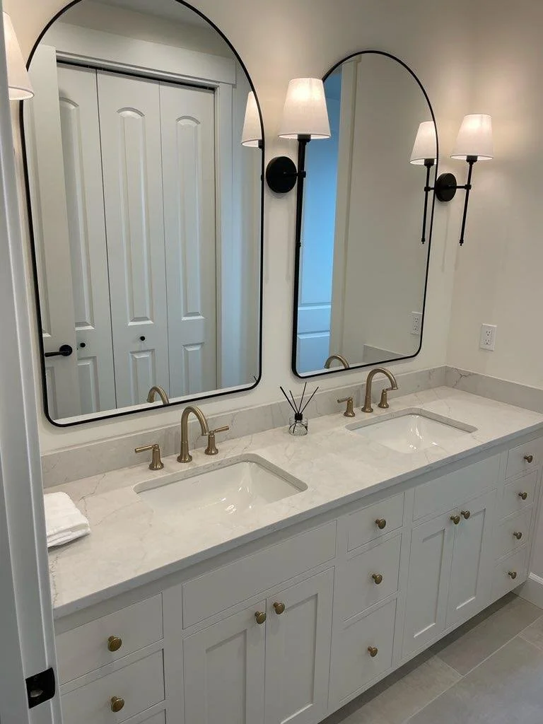 A bathroom vanity with two sinks, white marble countertop, gold faucets, and two large mirrors with black frames. There are two wall-mounted light fixtures with white shades between the mirrors. A folded towel and a small reed diffuser are on the cou