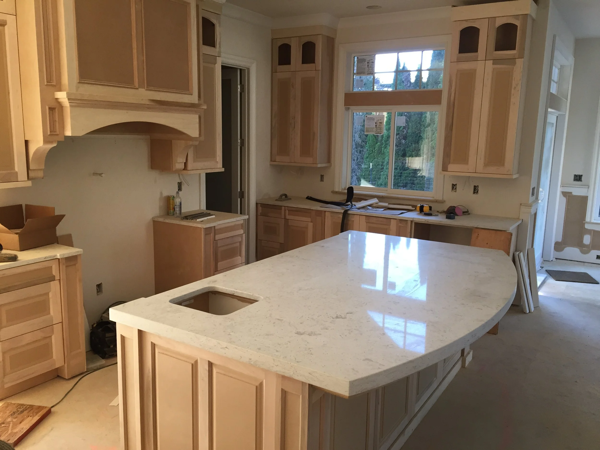 Unfinished kitchen with light wood cabinets, a large island with a white stone countertop, and window over the sink area. Construction tools and materials are present.