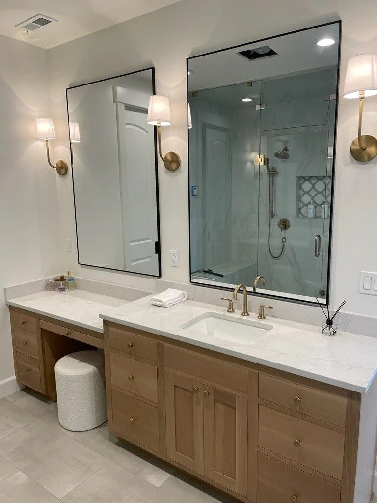 Modern bathroom vanity with a marble countertop, two large mirrors, and brass fixtures. Two wall-mounted lamps with white shades illuminate the area. A glass-enclosed shower with marble walls is visible in the mirror's reflection. There is a small wh