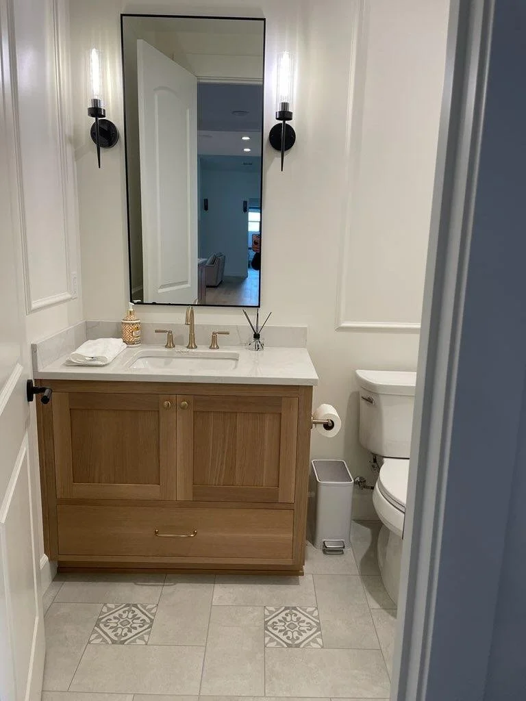 Small bathroom with a wooden vanity, white countertop, and rectangular mirror. Wall-mounted black light fixtures on either side of the mirror. Toilet with a silver toilet paper holder and trash can nearby. Tiled floor with decorative accent tiles.