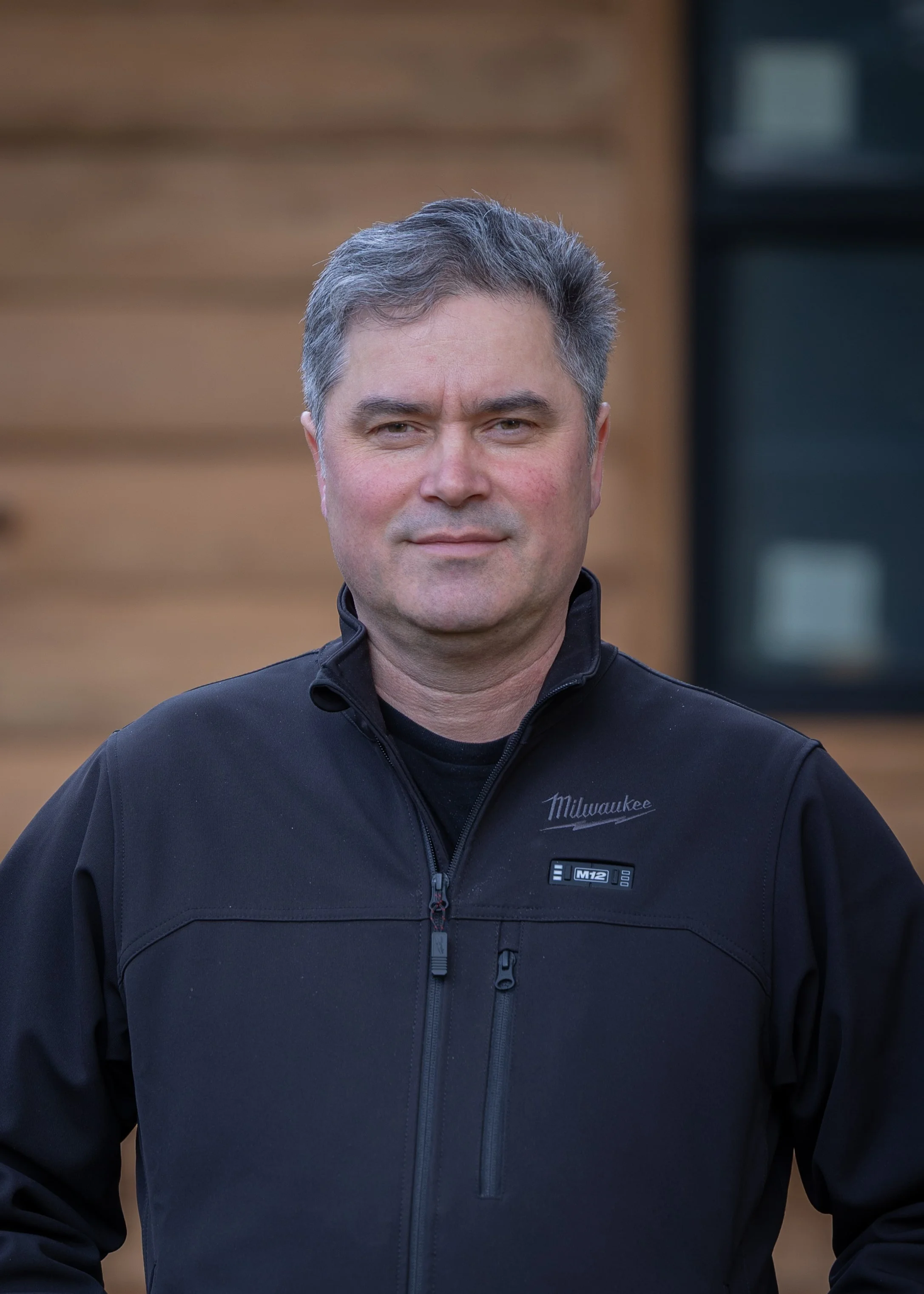 A middle-aged man with short, gray hair standing outdoors in front of a wooden building. He is wearing a black jacket with a zipper and a Milwaukee logo.