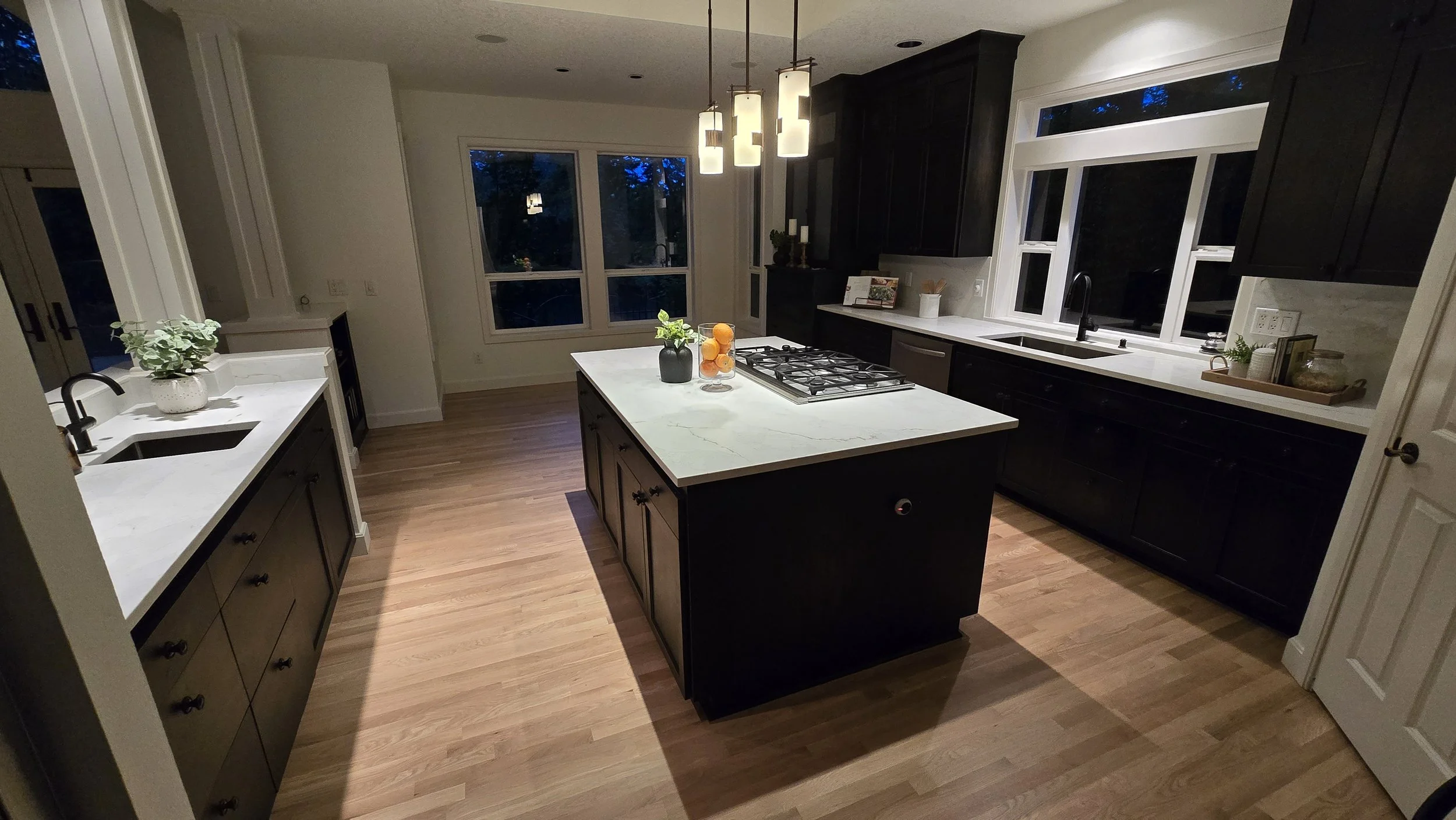 Modern kitchen with dark cabinets, white countertops, a kitchen island with a gas stove, wooden flooring, large windows, and a kitchen sink under the window.