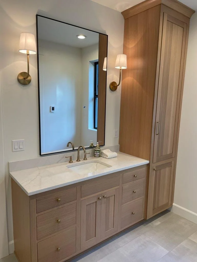 Bathroom vanity with a large mirror, two wall-mounted lamps, a beige wooden cabinet, and drawers, with a white stone countertop and a brass faucet.