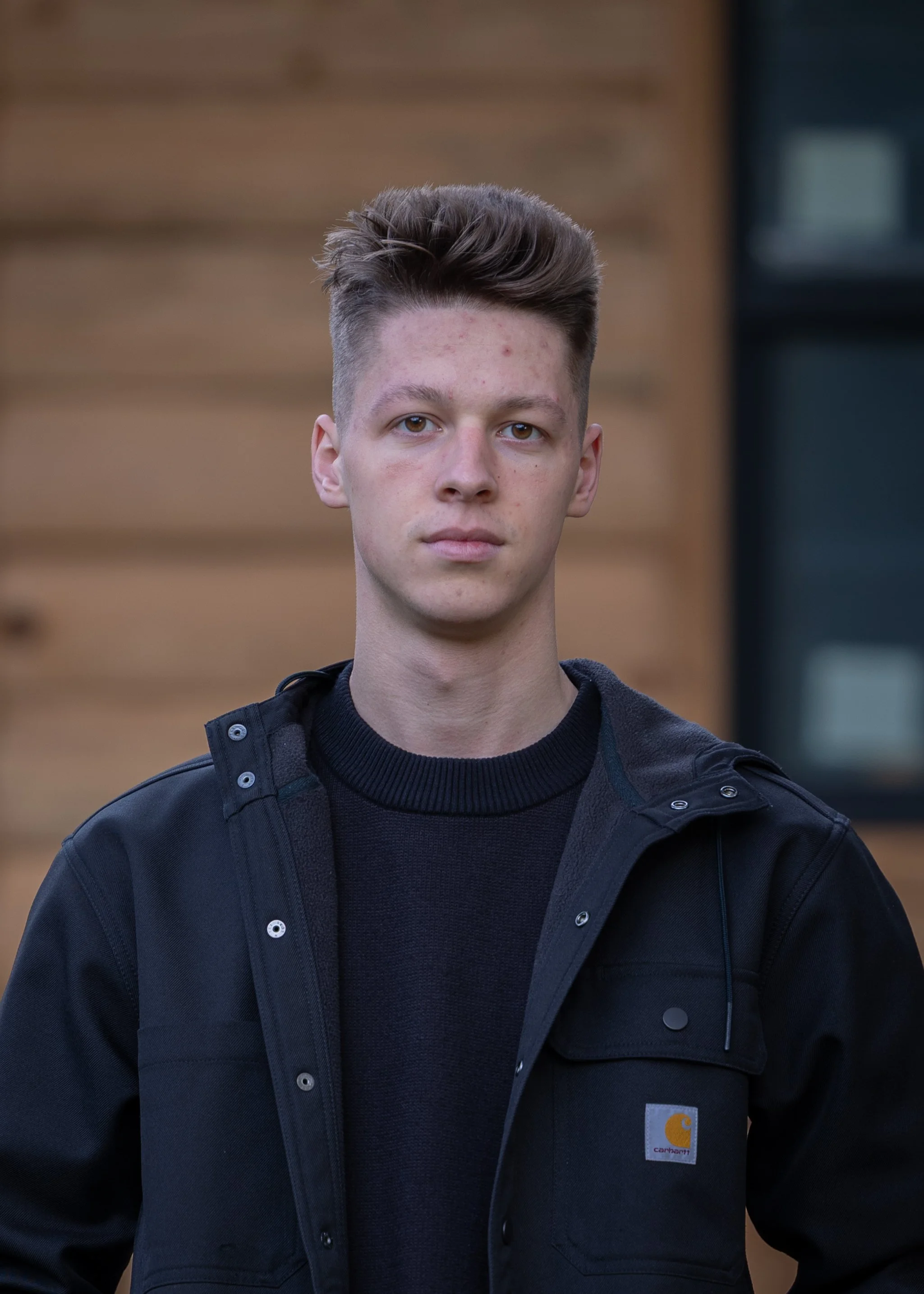A young man with styled brown hair, wearing a black Carhartt jacket and a black shirt, standing outdoors in front of a blurred wooden building background.