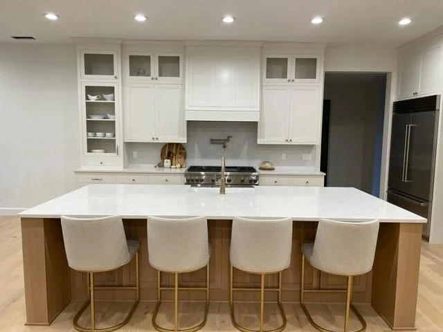 Modern kitchen with white cabinets, a large island with a white countertop, four beige barstools, stainless steel appliances, and a light wood floor.