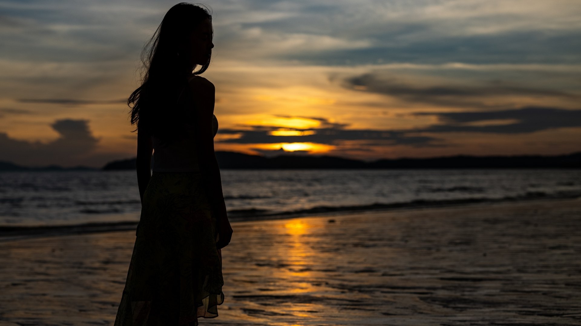 Silhouette of a woman standing on the beach during sunset with the ocean and cloudy sky in the background.