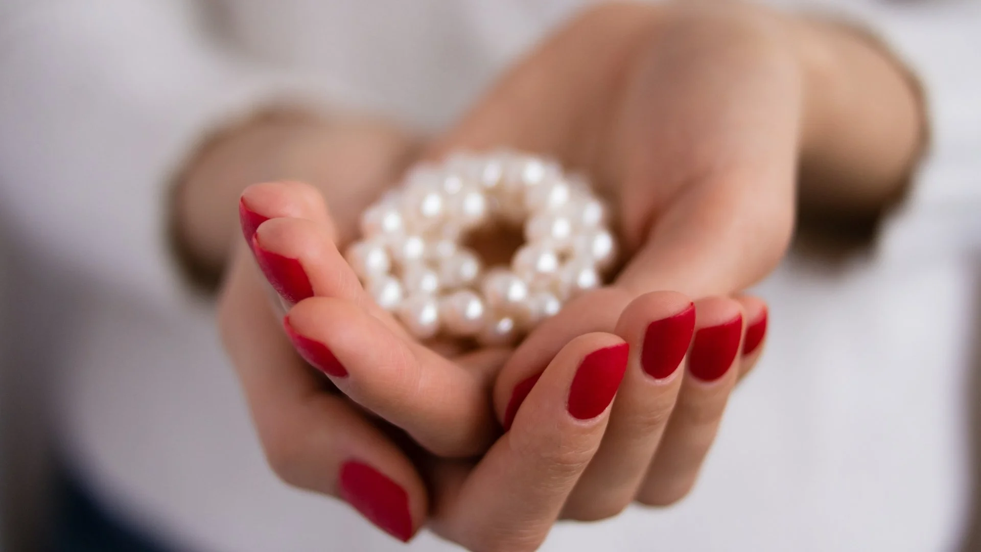 A hand with red-painted nails holding a circular pearl bracelet.