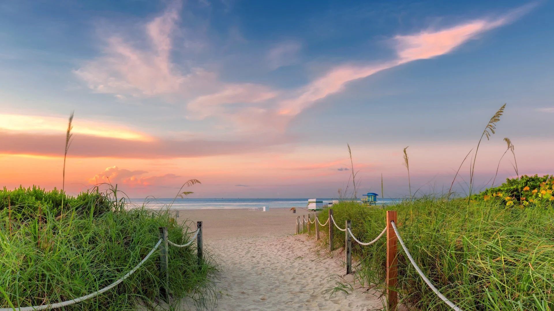 A sandy beach path lined with green bushes and tall grass leading to the ocean, under a colorful sunset sky with pink and blue clouds, and a blue lifeguard tower in the distance.