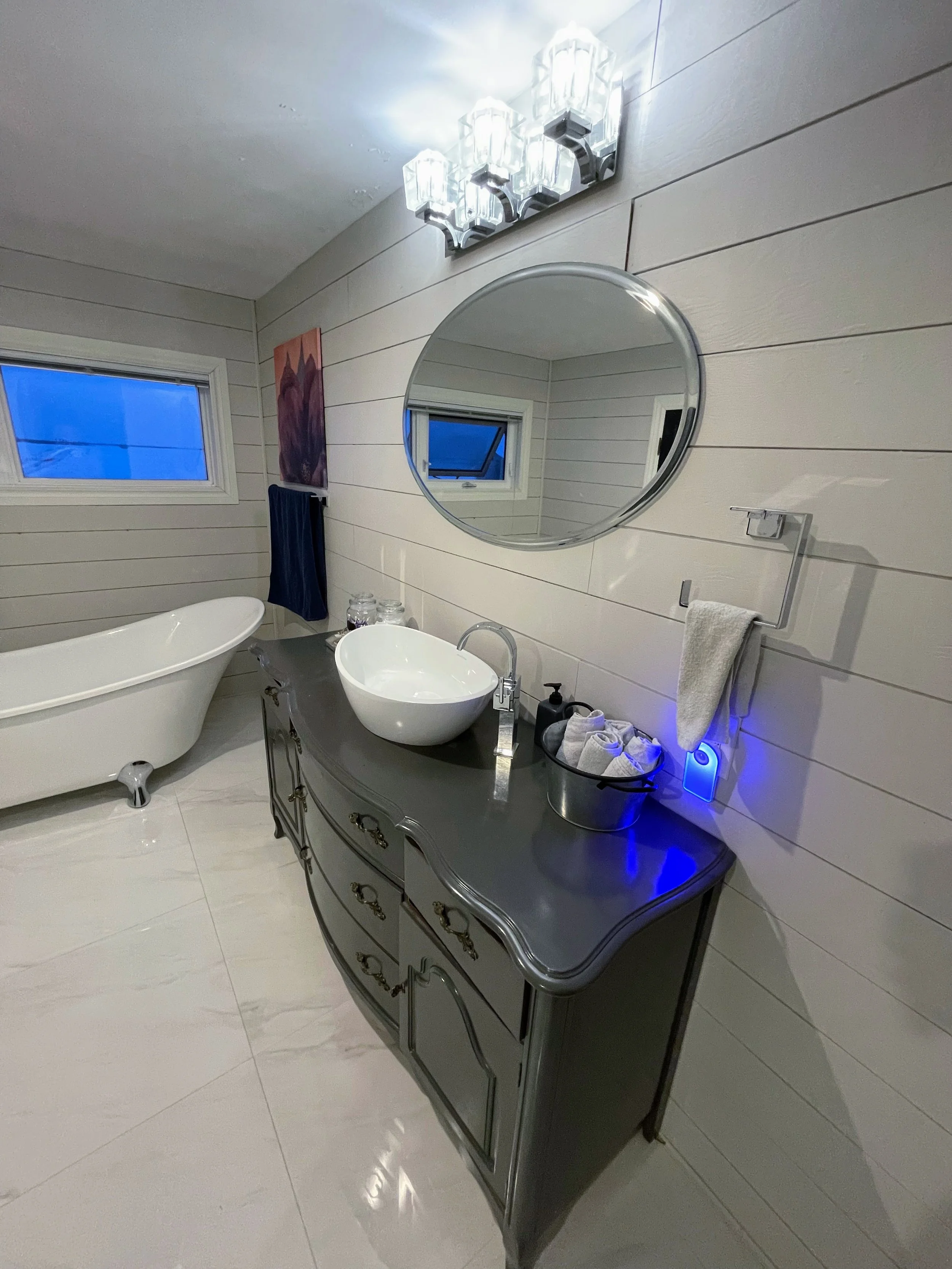 A modern bathroom with white wooden panel walls, a black vanity with a white vessel sink, a round mirror, a bathtub, a small frosted window, and a decorative washing area.