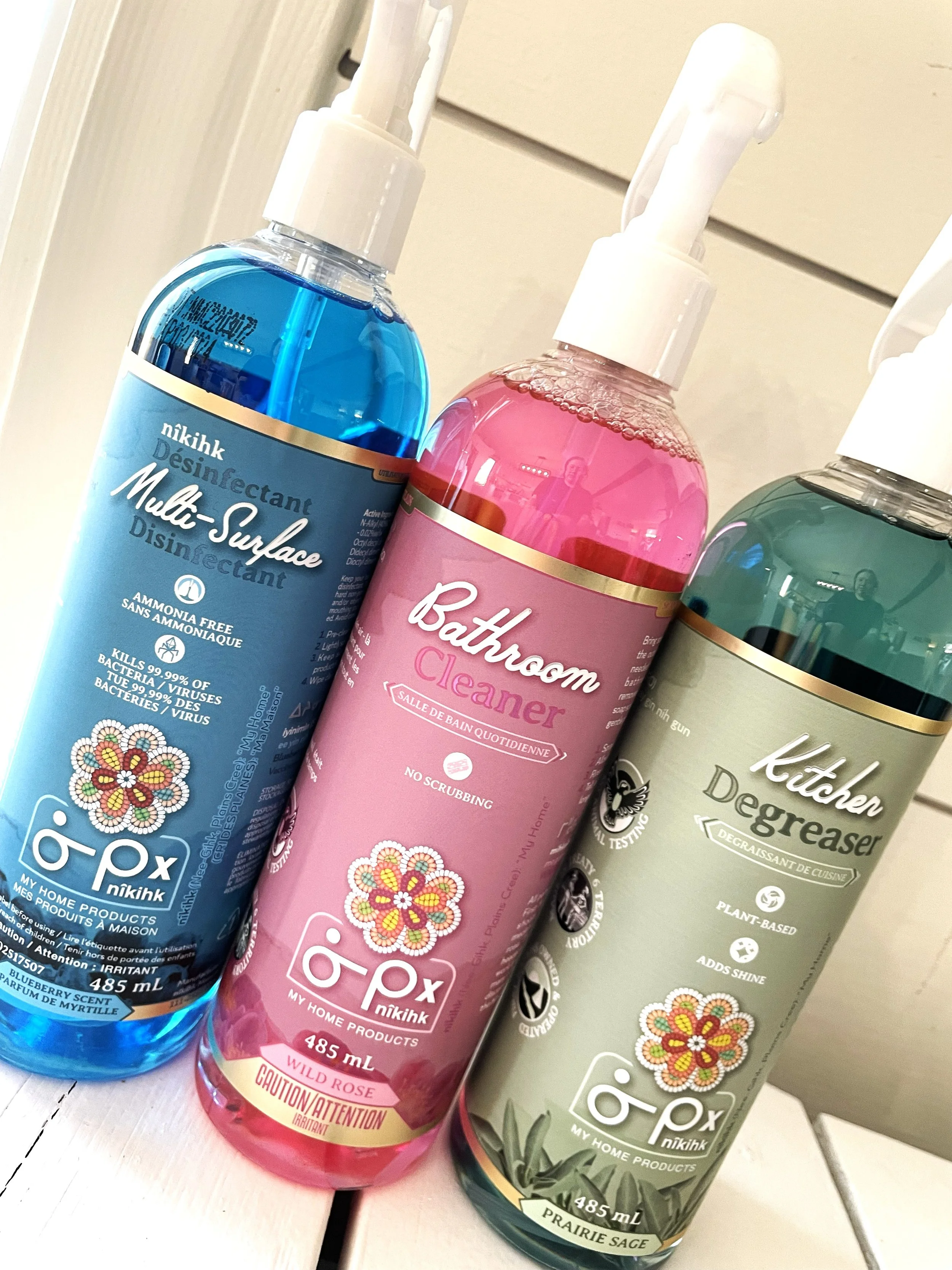 Three bottles of home cleaning products, including multi-surface disinfectant, bathroom cleaner, and kitchen degreaser, arranged on a white wooden surface.