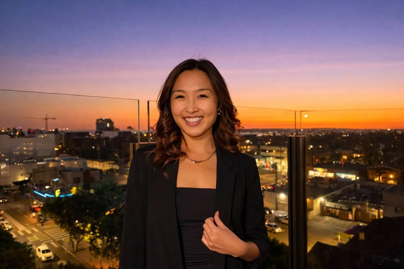 A woman smiling in a black blazer and dress standing on a rooftop with a sunset cityscape in the background.
