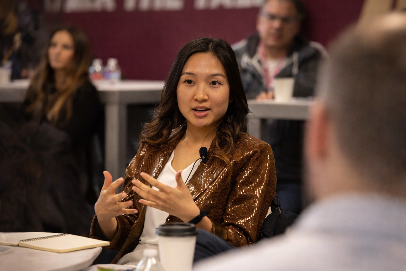 A woman speaking at a panel discussion, gesturing with her hands, wearing a sparkling brown jacket, with other attendees seated around her.