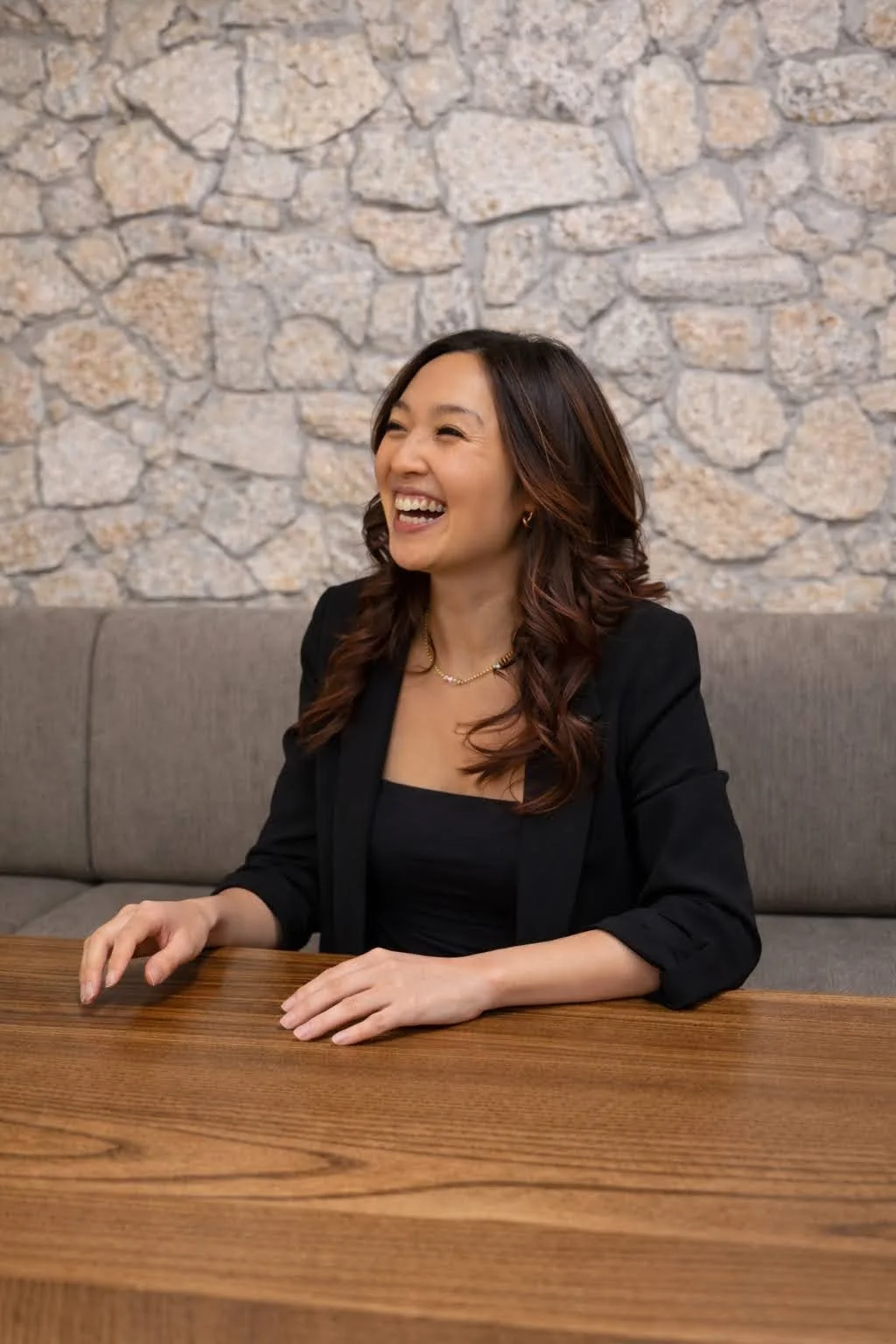 A smiling woman with wavy, dark brown hair sitting at a wooden table against a stone wall background.