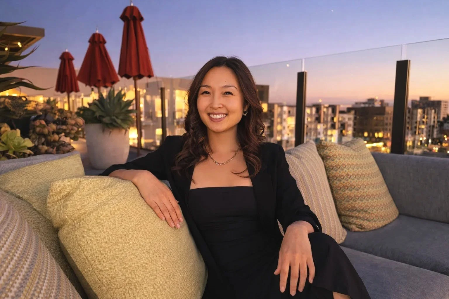 A woman smiling and sitting on a sofa on a rooftop terrace with city buildings in the background during sunset.