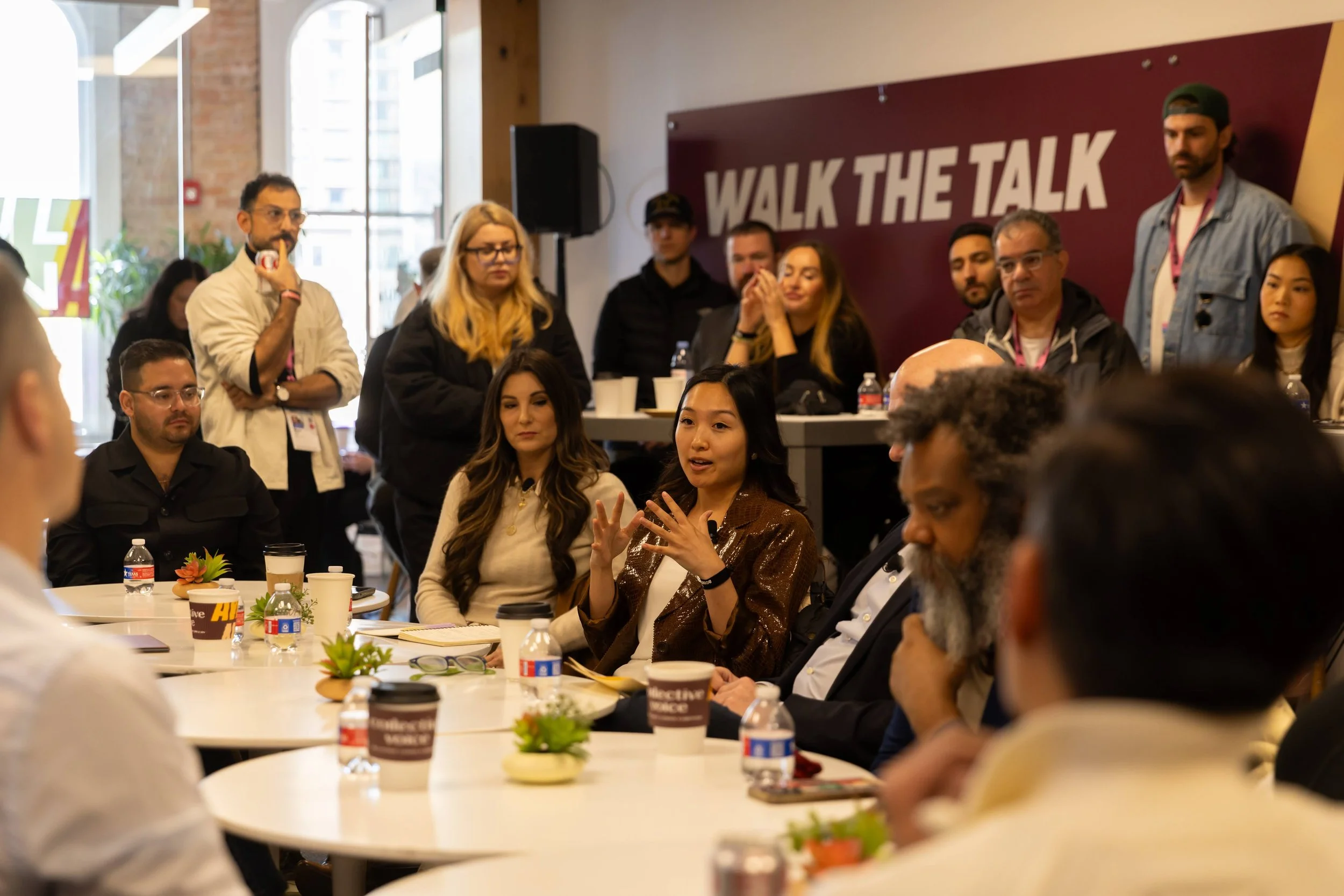 A diverse group of people attending a conference or workshop in a modern, urban venue with large windows and exposed brick walls. Some individuals are speaking, listening, or engaging in discussion. The background features a banner with the reading "WALK THE TALK." There are tables with refreshments and potted plants.