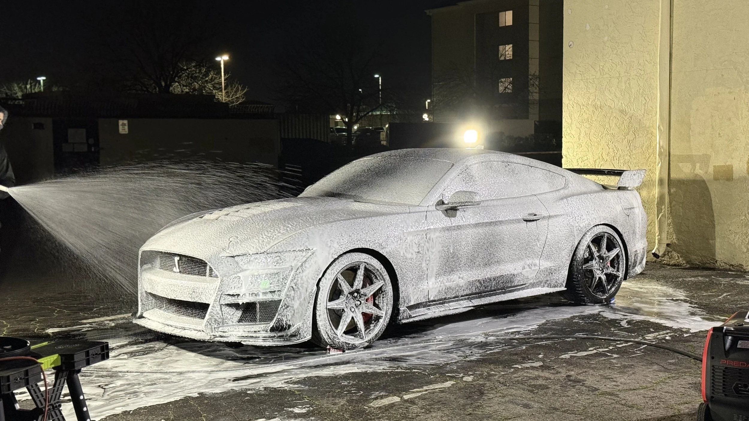 A silver sports car being washed with foam at night, with a person using a pressure washer on the left side.