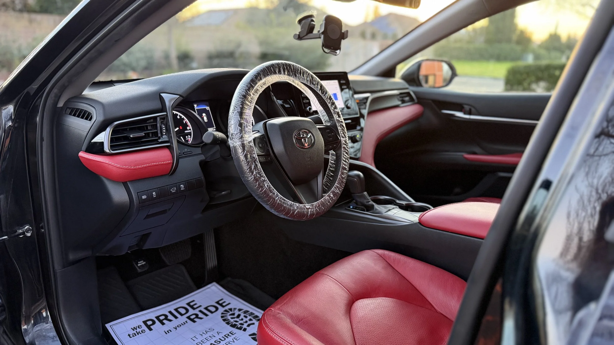 Interior of a Toyota vehicle with red seats and dashboard accents, plastic steering wheel cover, and a mounted phone holder visible, with a view outside through the window of a green landscape and sunset sky.
