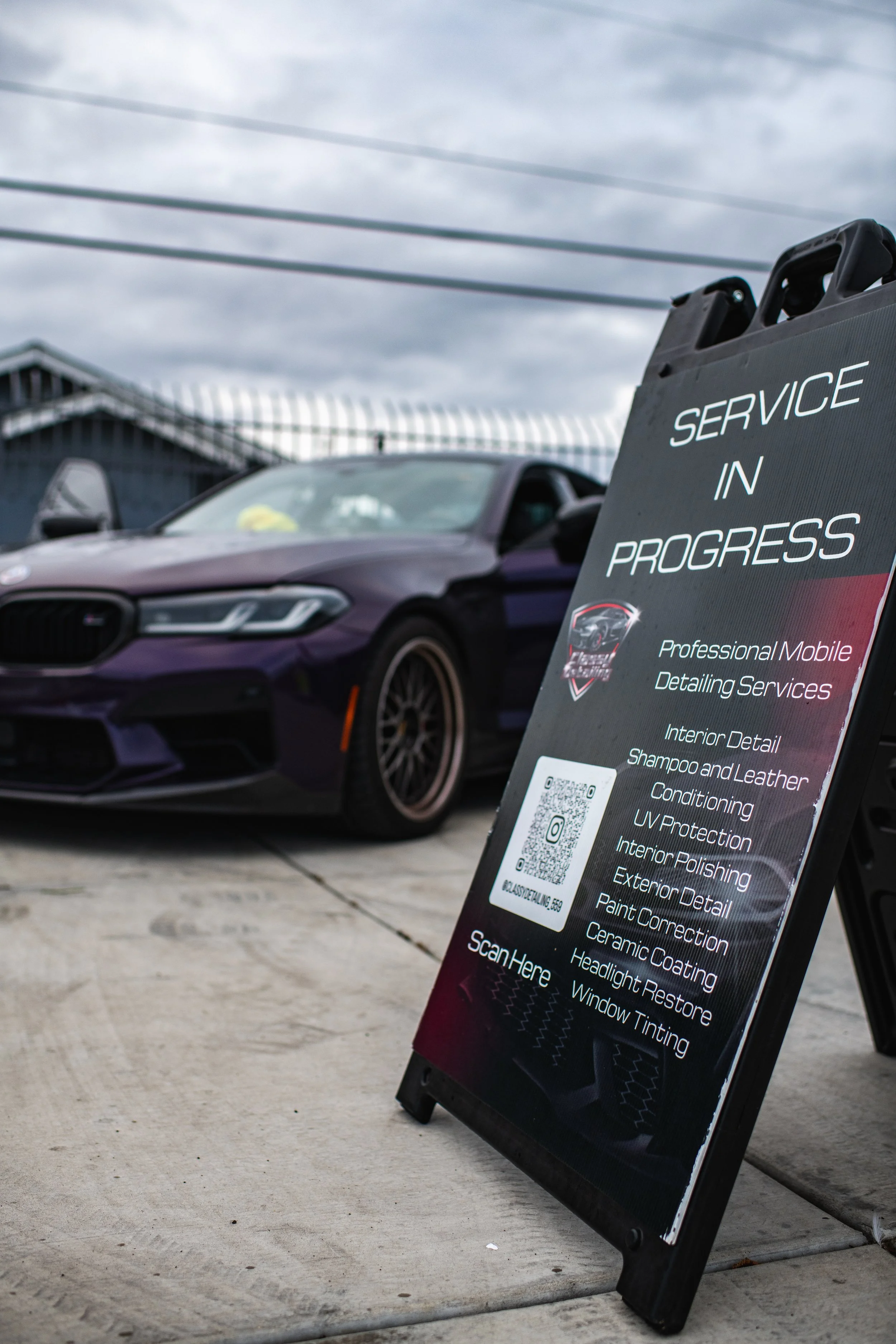 A purple car parked on a concrete surface and a service signboard in the foreground. The sign indicates ongoing vehicle detailing services.