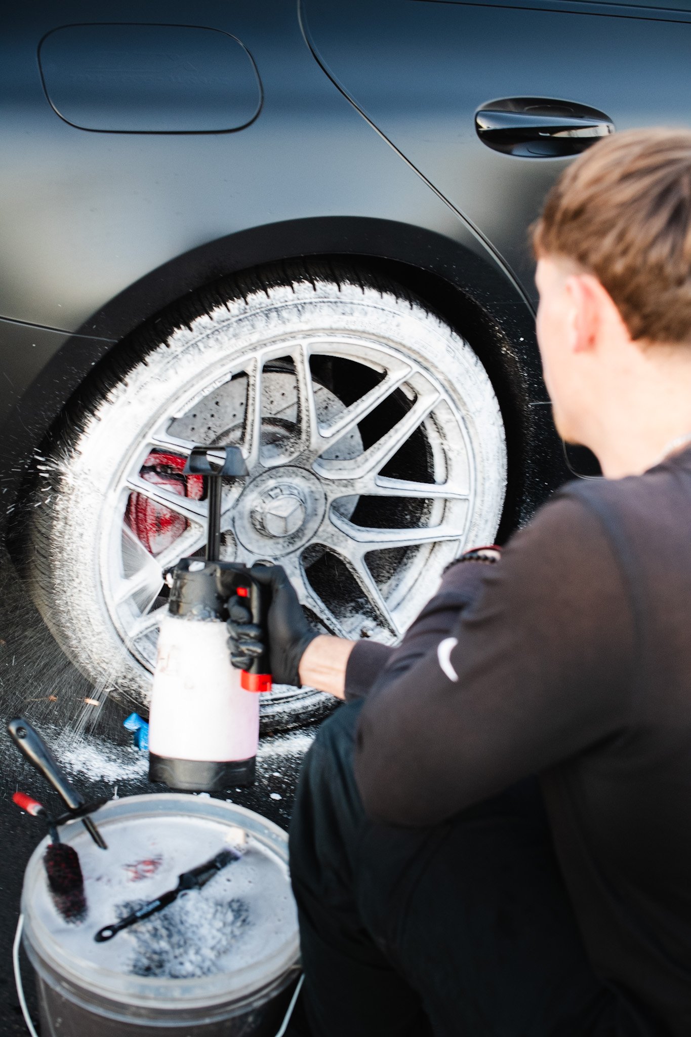 Man cleaning a black car's wheel with foam, using a spray bottle, brushes, and cleaning tools.
