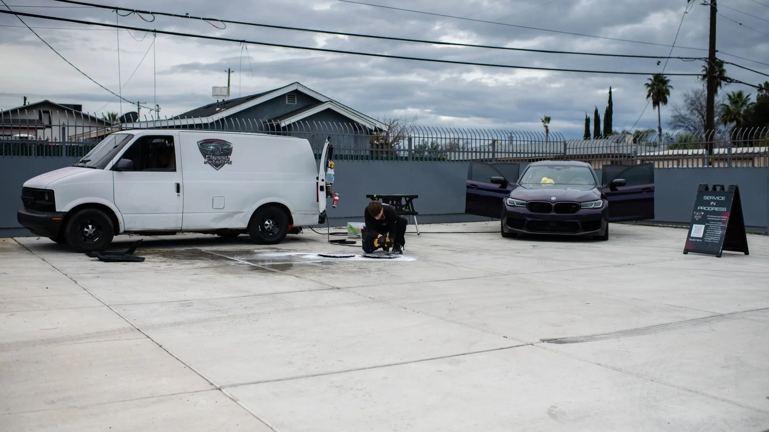 A person detailing a purple BMW with open doors in an outdoor lot under a cloudy sky with their white mobile detail van.