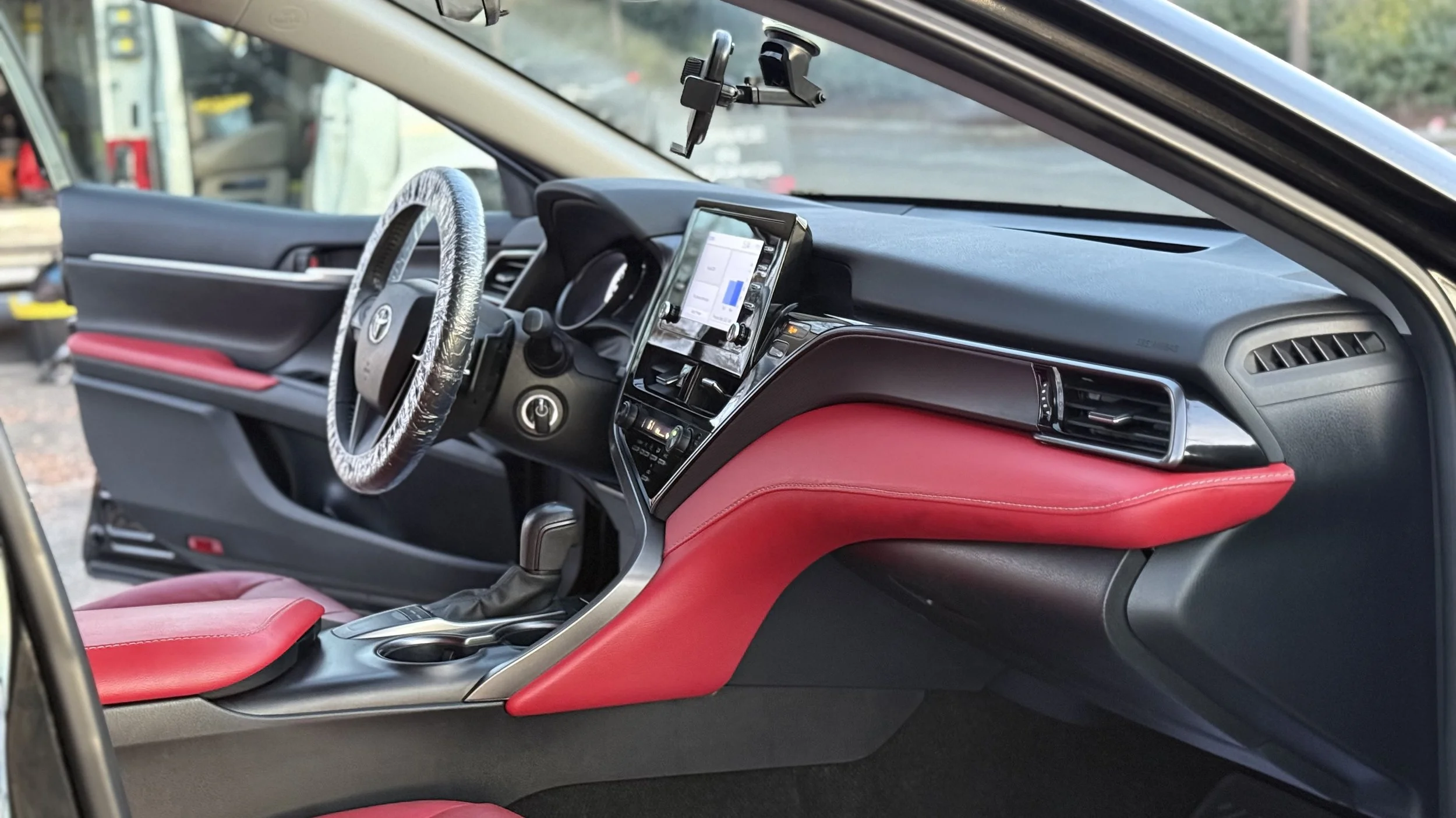 The interior of a modern vehicle featuring a dashboard, steering wheel with cover, touchscreen display, gear shift, and red and black upholstery.