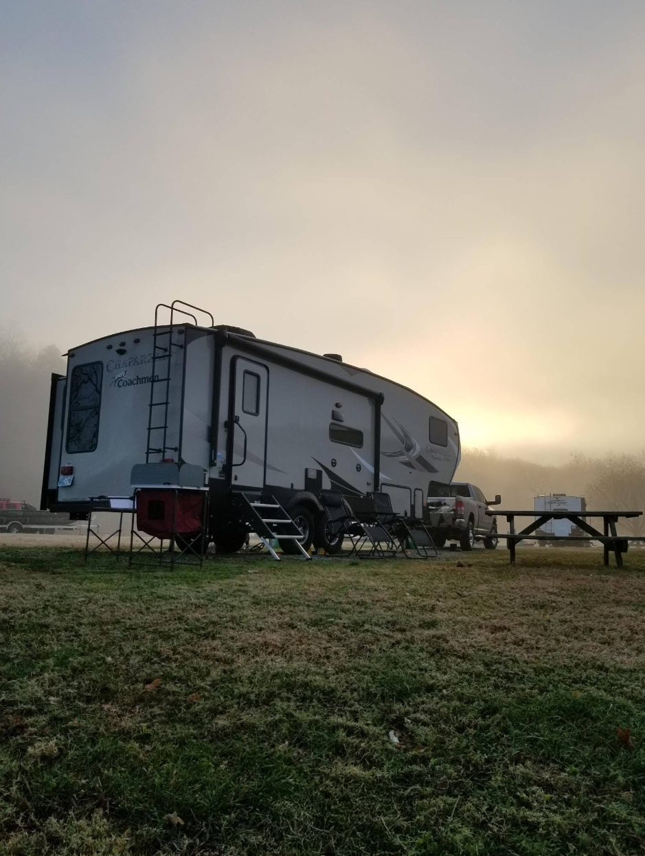 A recreational vehicle (RV) parked on a grassy area with outdoor chairs, a table, and a ladder, under an overcast sky with a bright spot near the horizon.