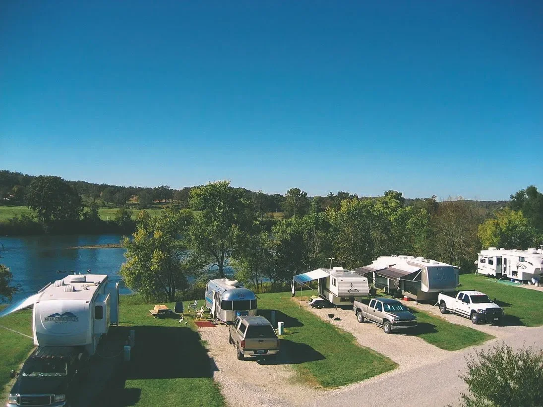 Campground by a river with multiple RVs and trucks parked on grassy and gravel areas, surrounded by trees and hills under a clear blue sky.