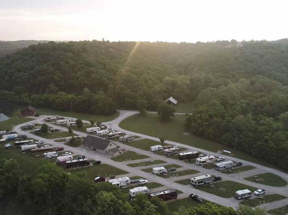 Aerial view of a campground with multiple RVs and trailers parked along gravel roads, surrounded by green hills and trees, with a small building in the center.