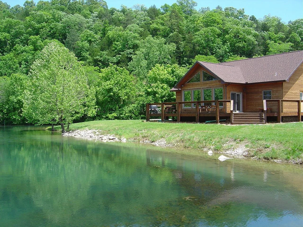 A wooden house with a deck beside a peaceful green lake, surrounded by lush trees and forested hills in the background.