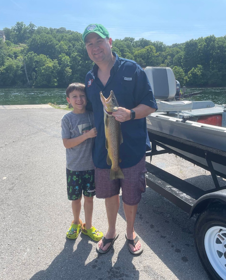 A man and a boy are standing outdoors near a body of water, holding a large fish they caught. The man is wearing a navy blue shirt, plaid shorts, flip-flops, and a green cap with a logo. The boy is wearing a gray T-shirt, black shorts with colorful designs, and bright green Crocs. There is a boat on a trailer behind them, and the background shows trees and a clear blue sky.
