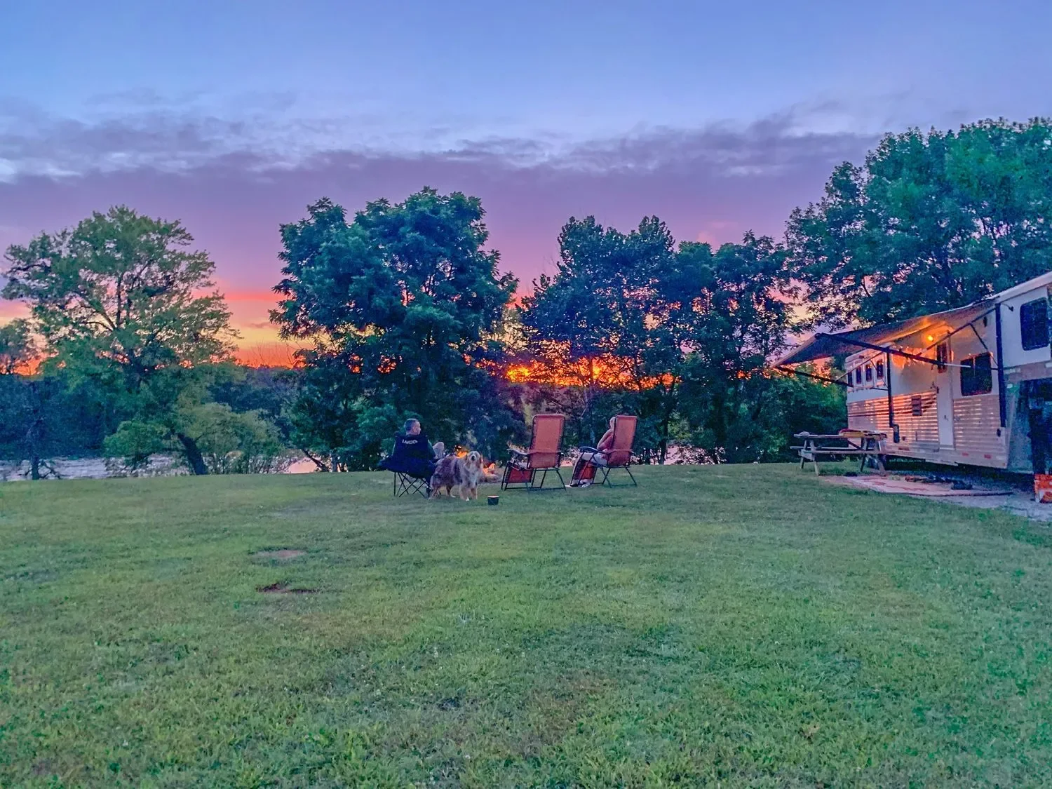 People sitting outside near a camper trailer at sunset with a dog, surrounded by trees and a grassy field.
