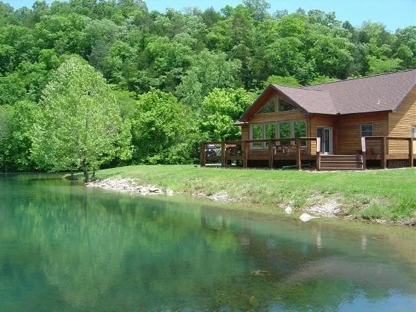 A wooden cabin with a deck beside a lake surrounded by lush green trees and a grassy area.