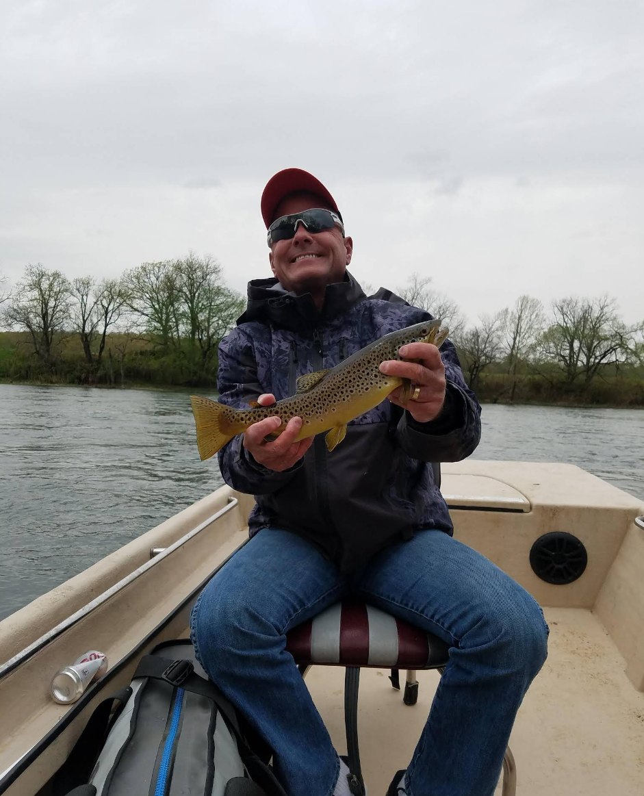 Man in a boat holding a fishing caught fish, wearing sunglasses and a red cap with a cloudy sky and bare trees in the background.