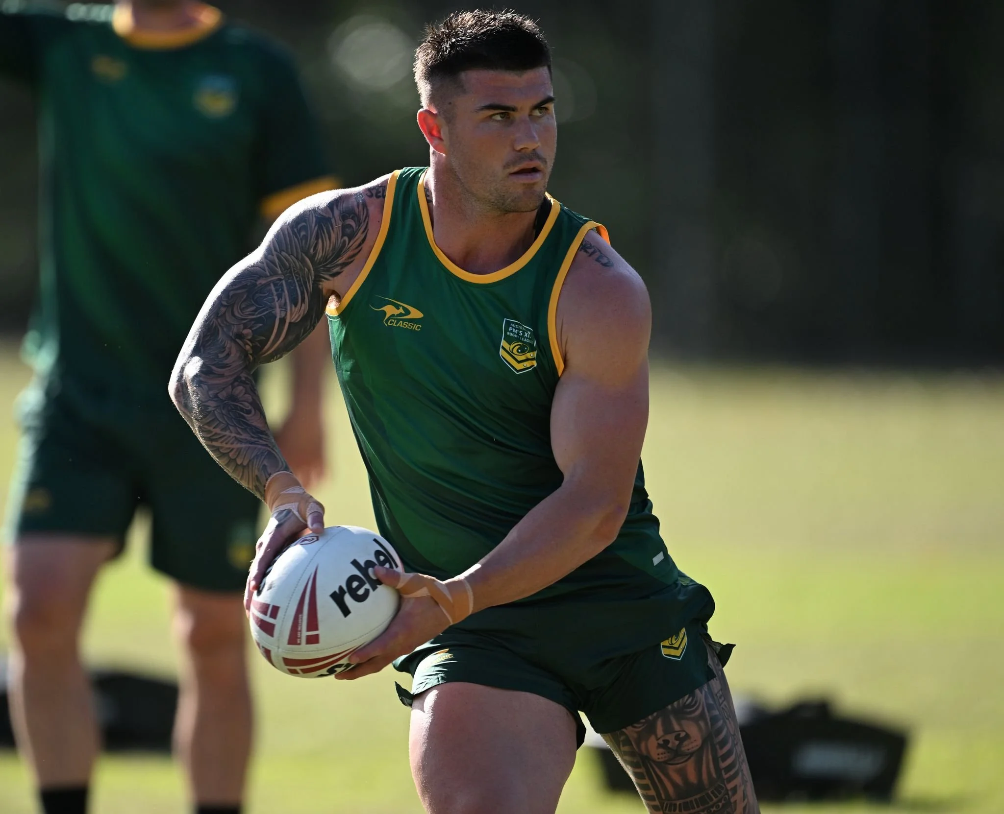 A male athlete holding a ball during a sports practice or game, wearing a green and yellow sleeveless uniform with tattoos on his arm, on a grassy field.