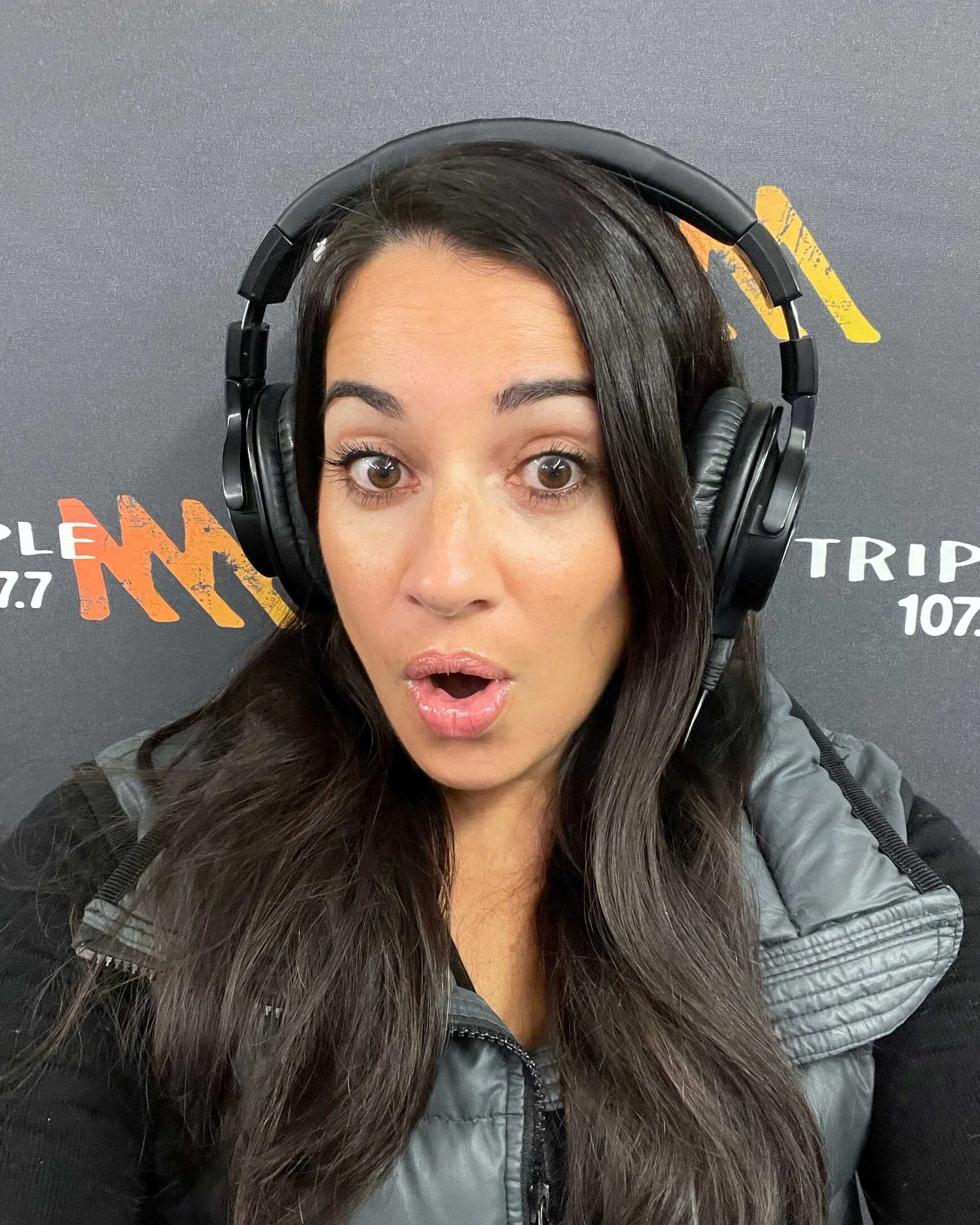 Close-up of a woman with long dark hair wearing a black jacket with a faux fur collar and large over-ear headphones, standing in front of a black background with orange and white text.