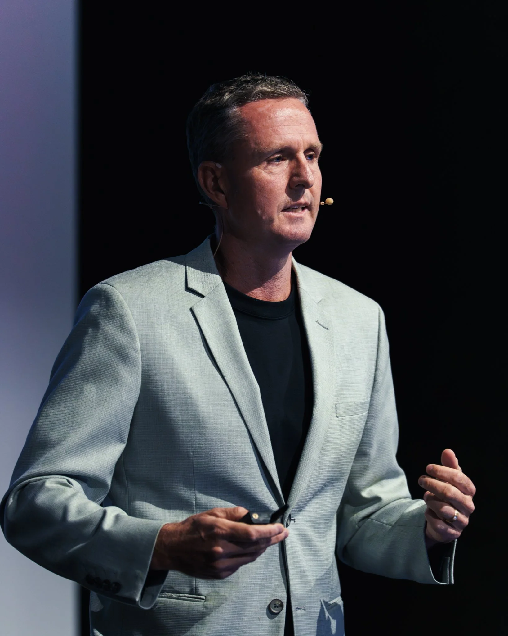 A man giving a presentation or speech, holding a remote control in his right hand, wearing a light gray blazer and black shirt, with a microphone headset, standing against a dark background.