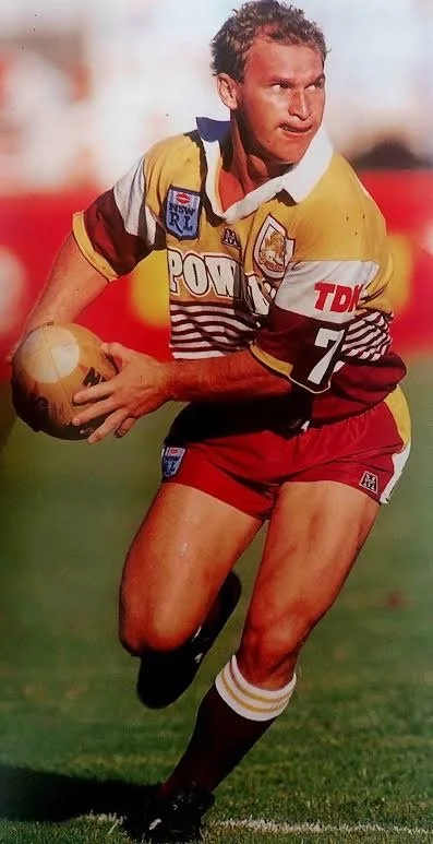 A rugby player in a yellow and maroon uniform holding a rugby ball, running on the field during a game.