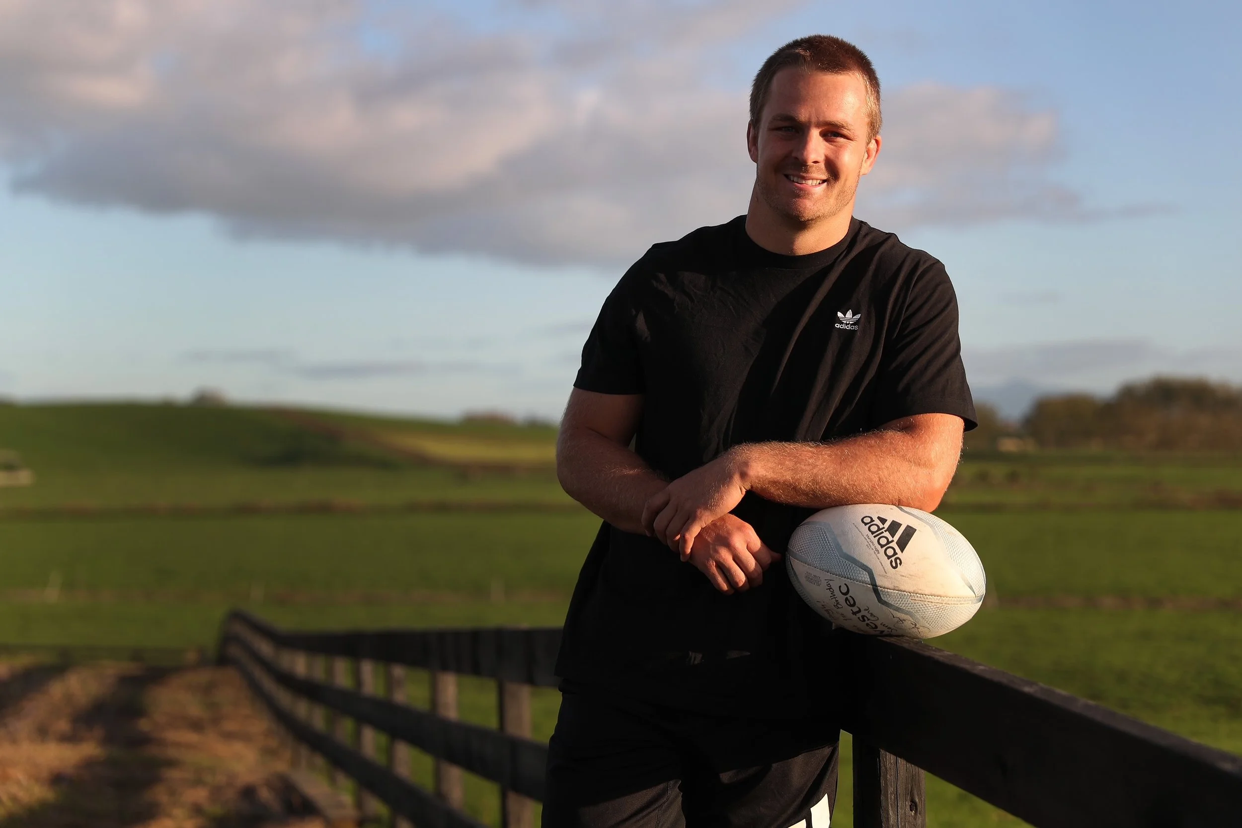 A young man in a black Adidas t-shirt holding a white Adidas soccer ball, standing outdoors near a wooden fence with a landscape of green fields and a cloudy sky in the background.
