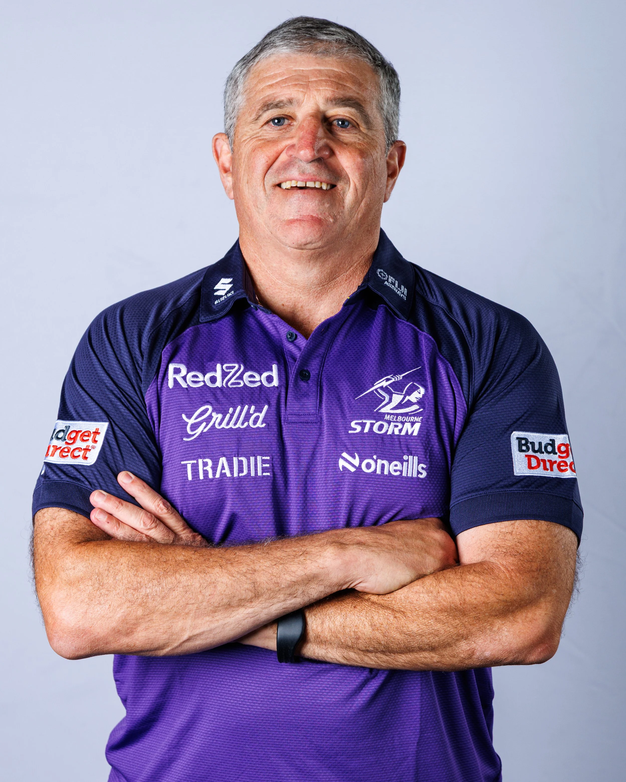 A middle-aged man with short gray hair and blue eyes, wearing a purple and navy Melbourne Storm rugby jersey with various logos, is standing with his arms crossed and smiling.