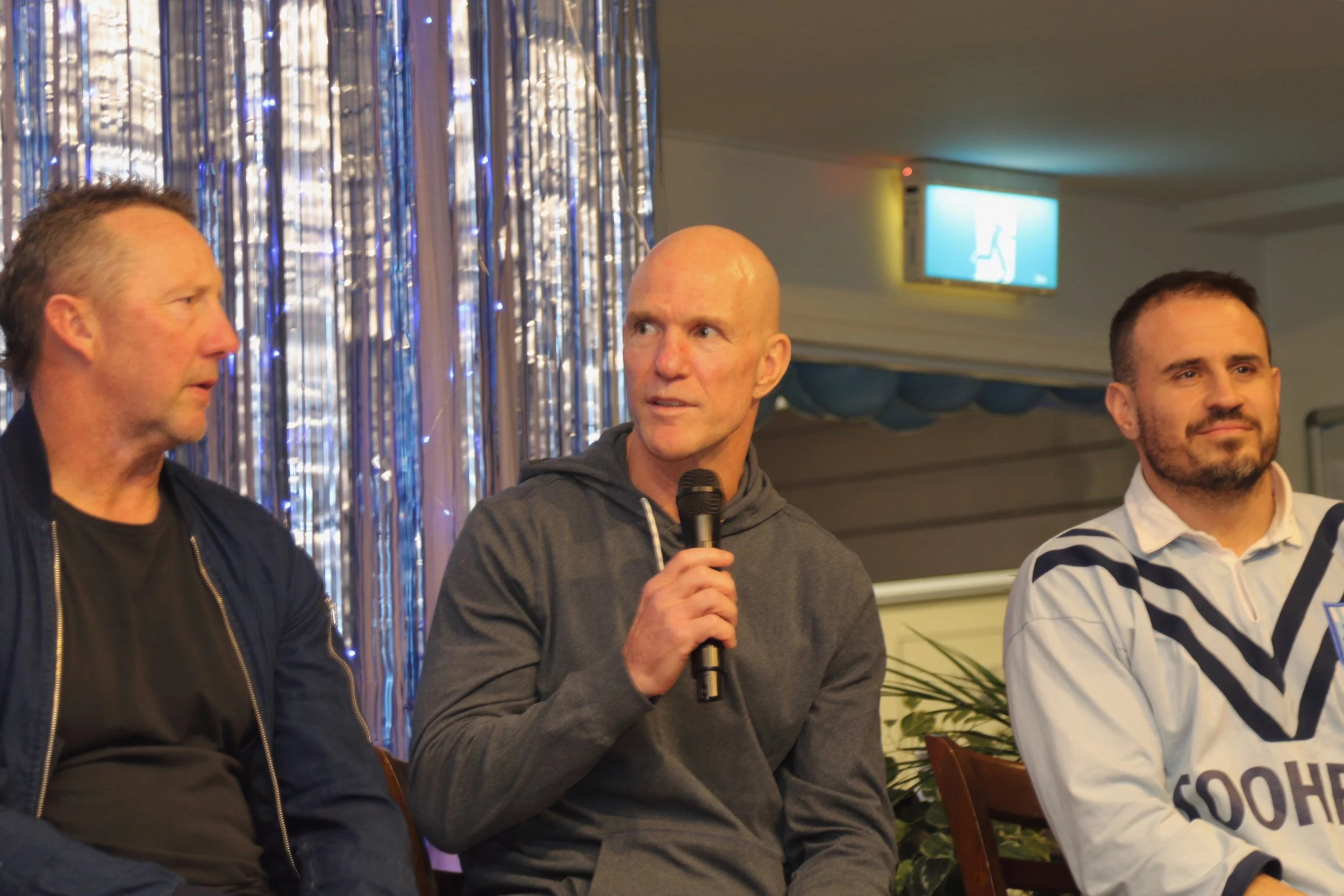Three men sitting at a panel discussion; the man in the middle is holding a microphone and appears to be speaking, with a backdrop of decorative curtains and a screen showing a logo.