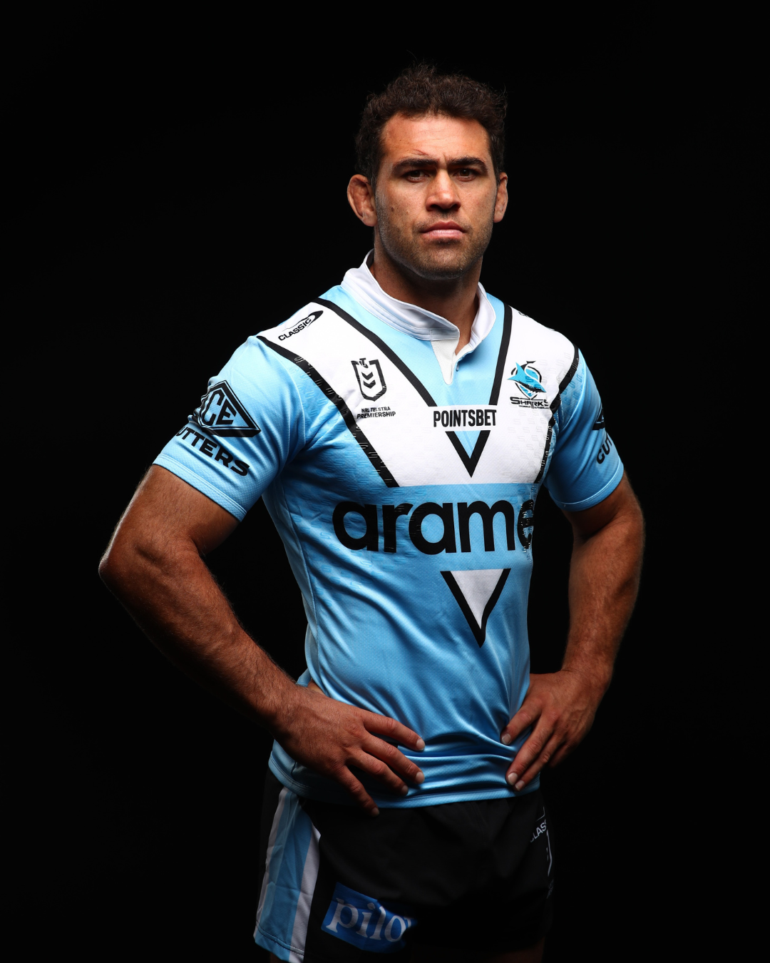 A rugby player wearing a light blue and white jersey with various sponsor logos, standing with hands on hips against a black background.