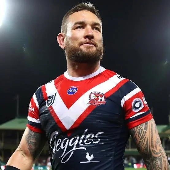 A rugby player in a red, white, and navy-blue uniform on a field at night, with tattoos on his arms and a short beard, looking slightly upward.