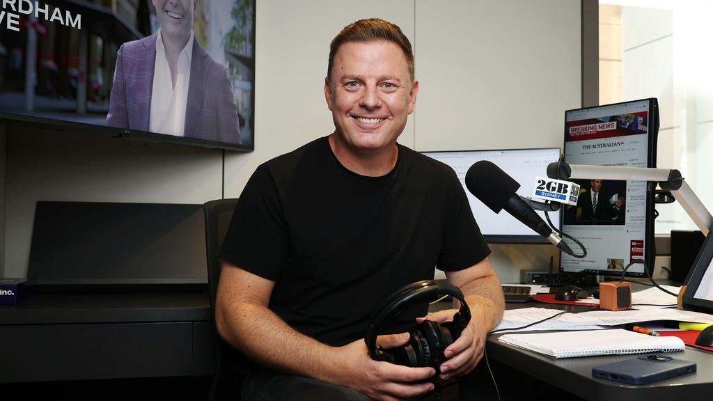 Man smiling, sitting at a desk in a radio studio, holding headphones, with microphones and computer monitors around him.