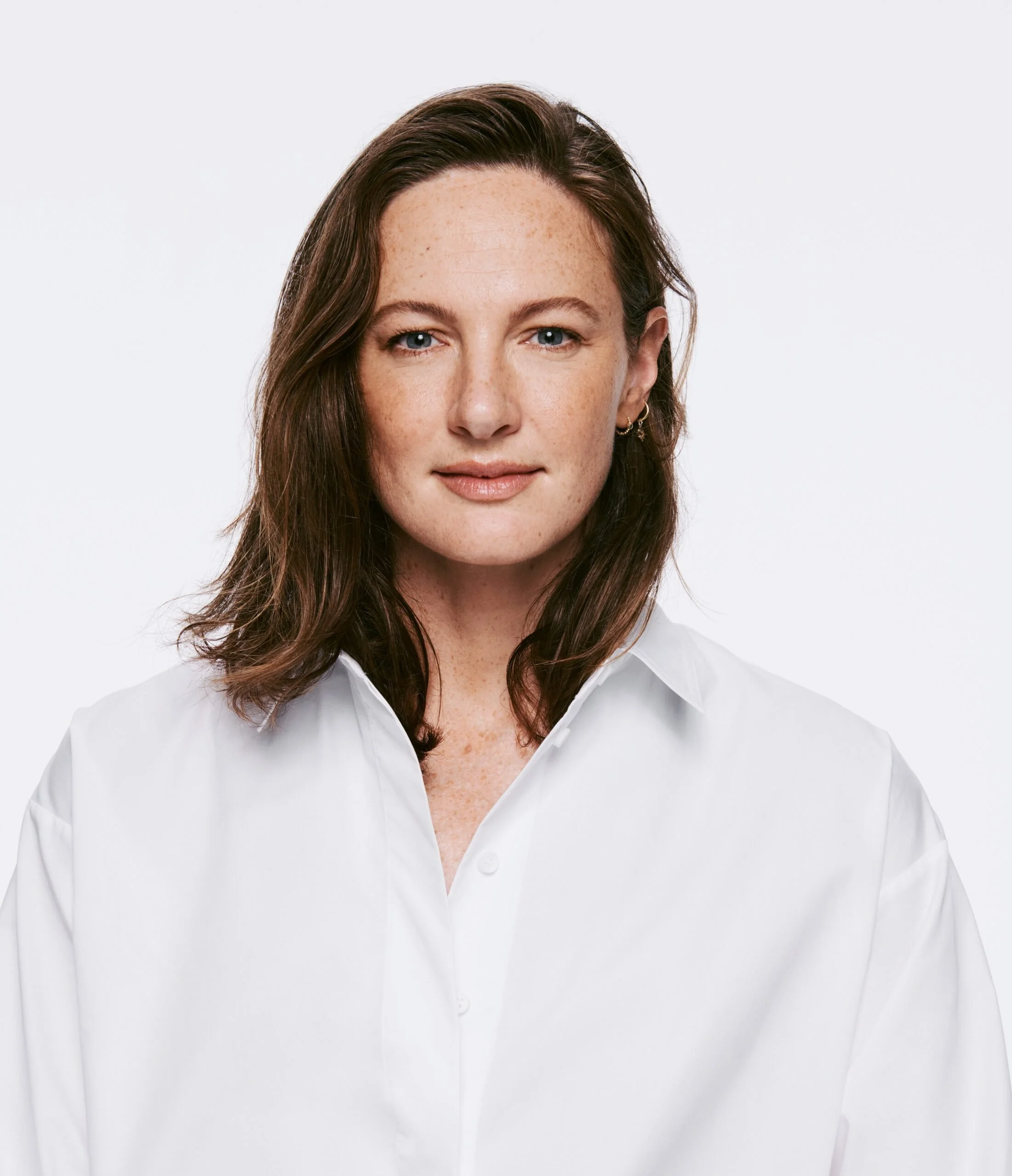 Close-up of a woman with medium-length brown hair, blue eyes, wearing a white button-up shirt, standing against a plain white background.
