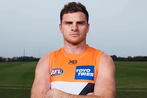 A male athlete in an orange and white sports jersey standing on a grassy field with arms crossed, looking at the camera.